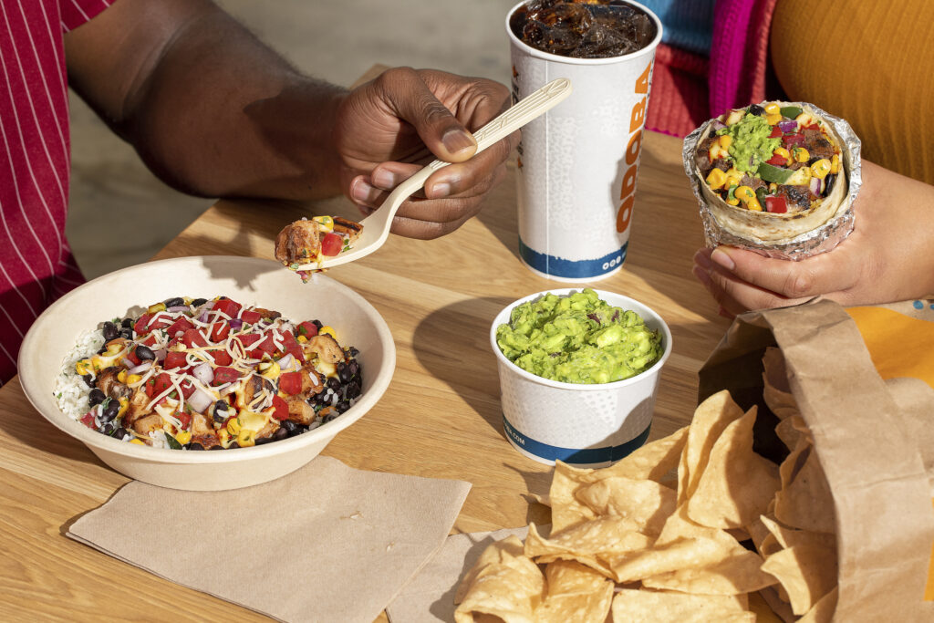 QDOBA meal with a burrito, rice bowl topped with beans, corn, cheese, and salsa, served with guacamole, tortilla chips, and a drink.