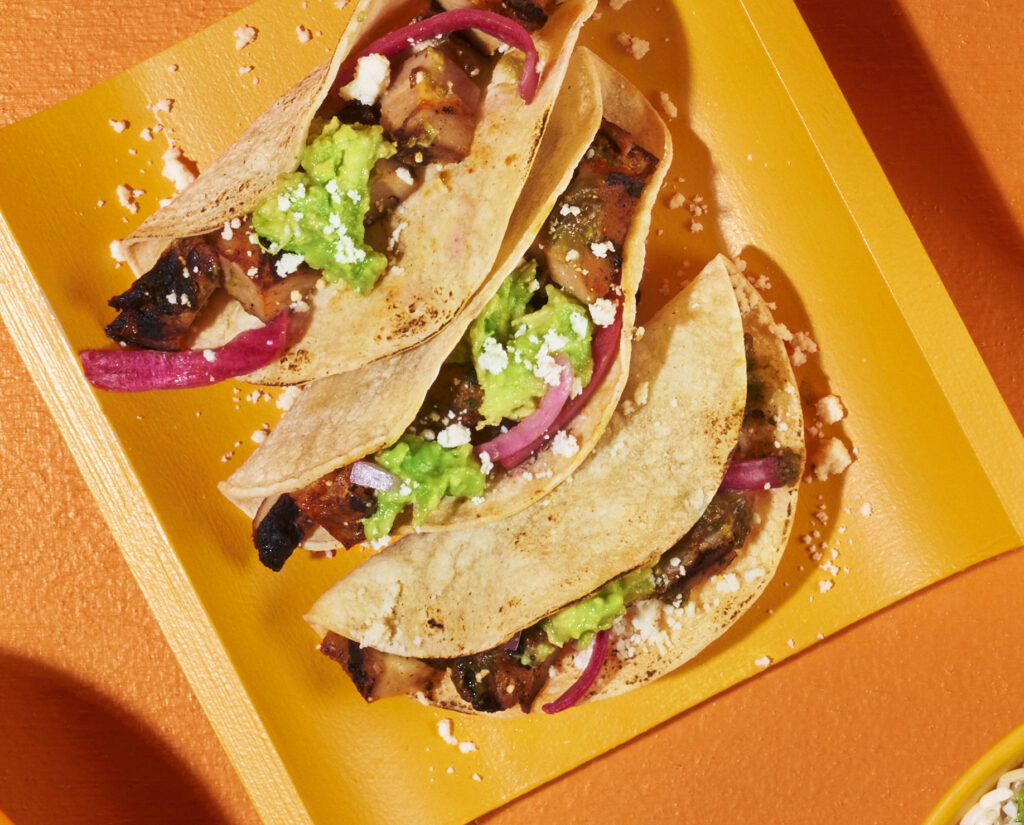 “Three QDOBA street tacos filled with grilled meat, guacamole, pickled red onions, and crumbled cheese on a bright orange tray.”
